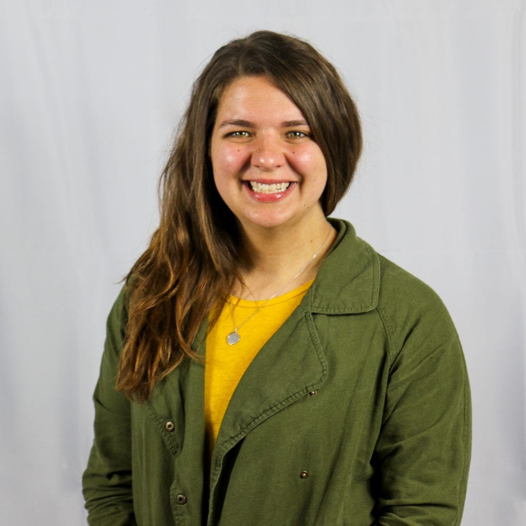 White female with long brown hair wearing a yellow shirt and green jacket.