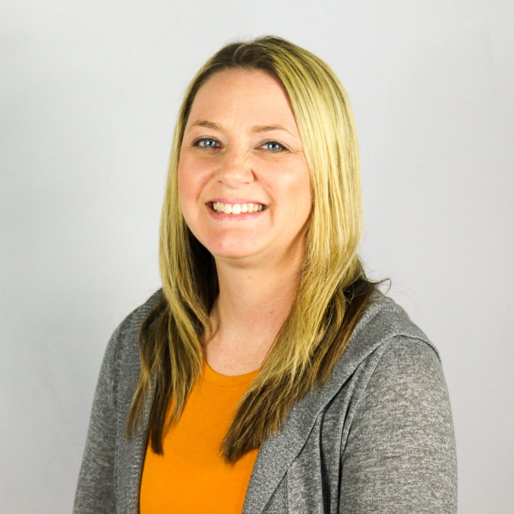 White female with long blonde hair and dark undertones, wearing an orange shirt with grey sweater.