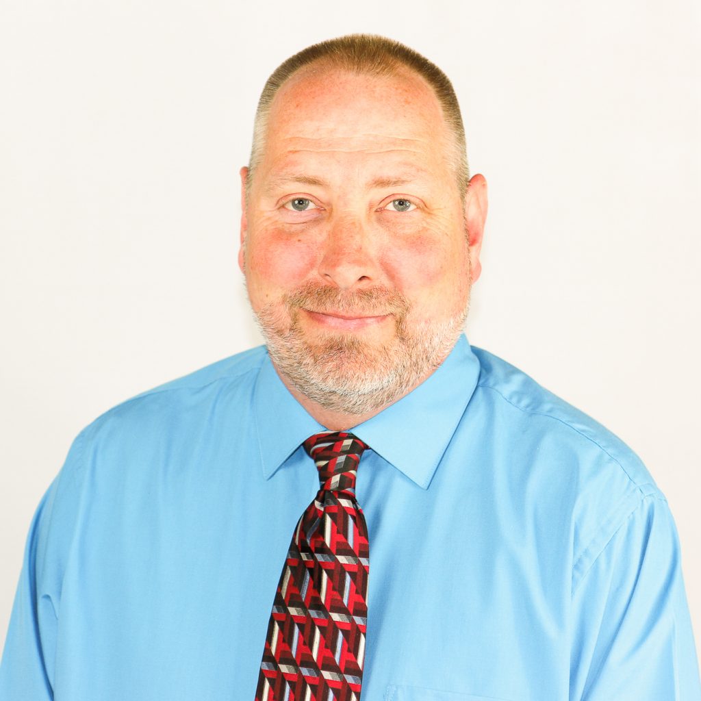 White male with short-cropped light brown hair, and a neatly groomed short beard in a blue shirt, and a patterned tie in shades of red, black, and white.