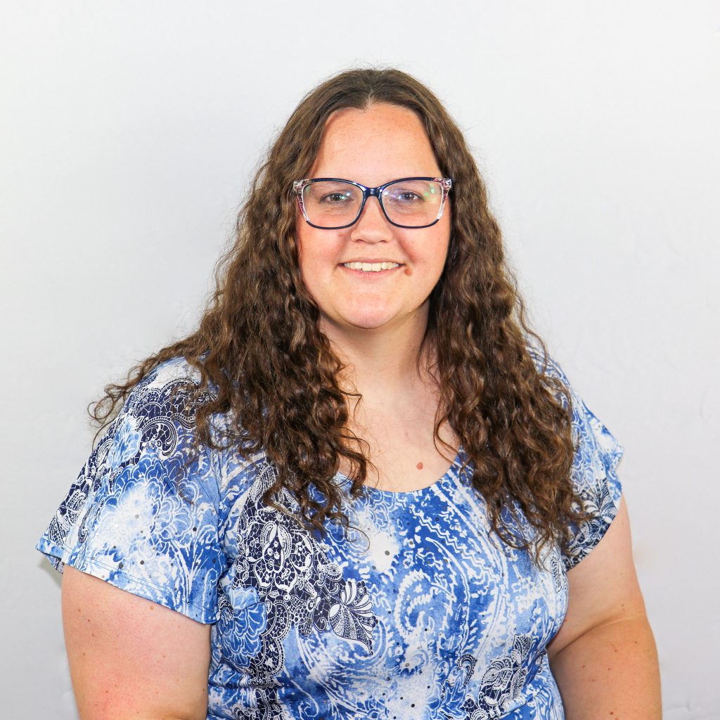 White female with long natural curly brown hair, rectangular eyeglasses and wearing a blue patterned blouse with a mic of light and dark tones.