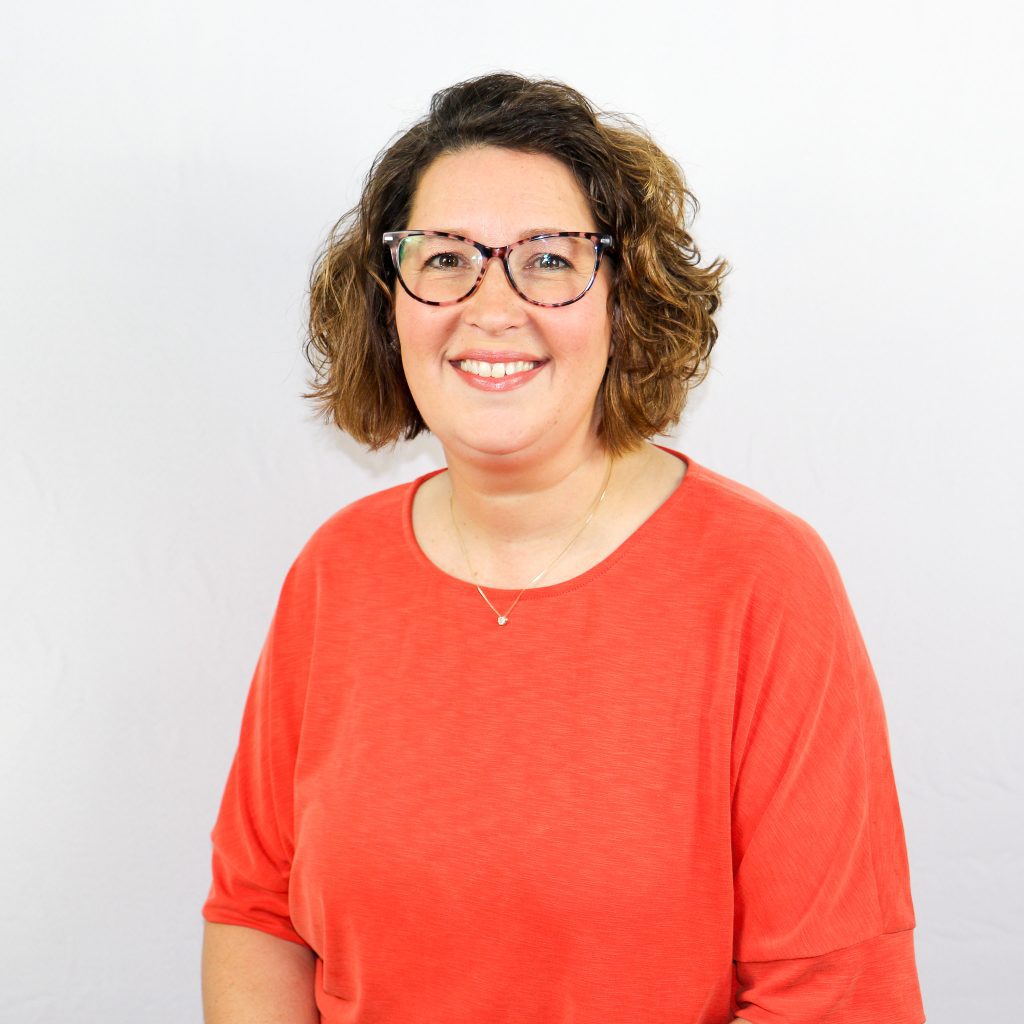 White female smiling with short wavy brown hair and glasses in an orange shirt.