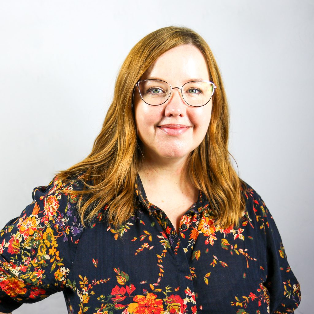 White female with shoulder-length wavy red hair wearing round glasses and a floral shirt.