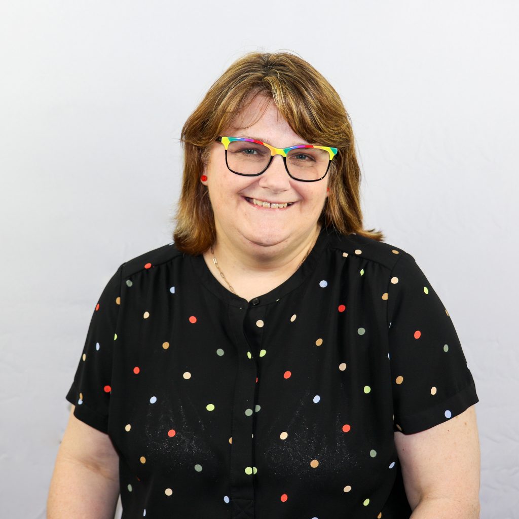White female wearing rainbow colored glasses with shoulder length reddish brown hair and black polka dot shirt