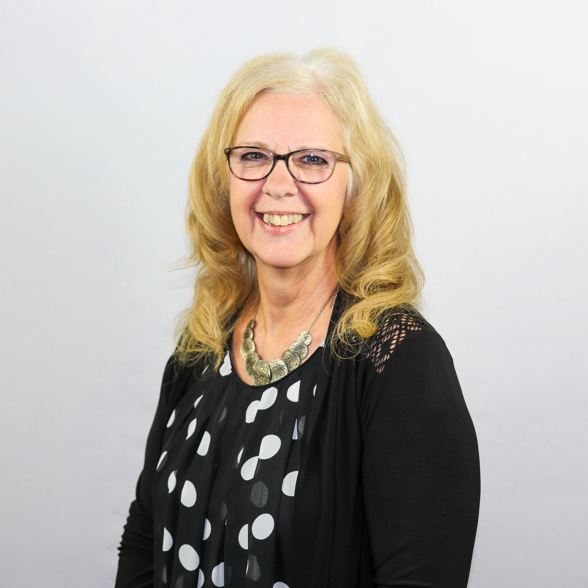 White female with curly shoulder length blonde hair wearing a black and white polka dot shirt.