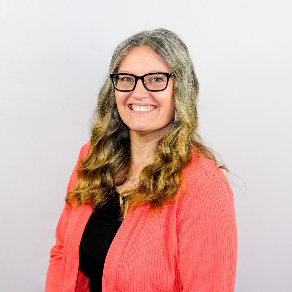 White female with wavy blonde hair wearing black rectangular eyeglasses, a black shirt and pink sweater.