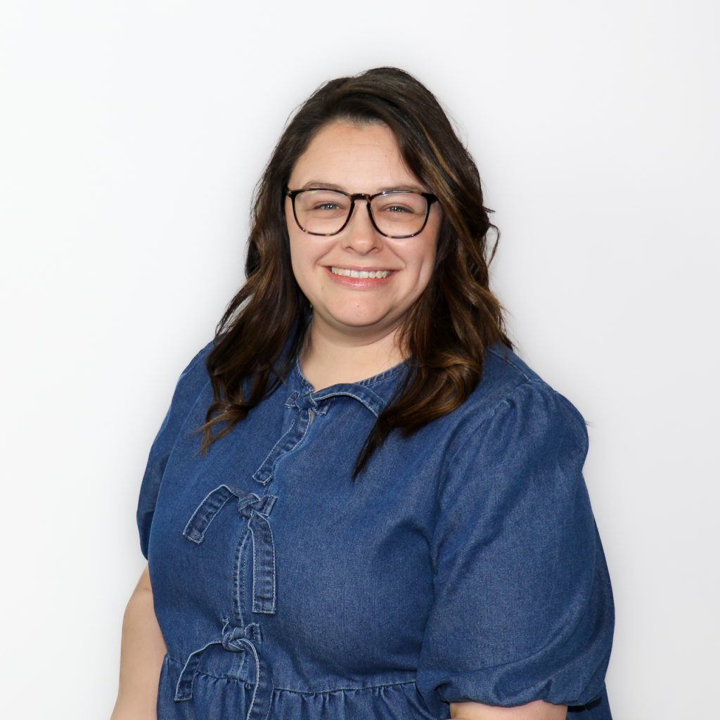 White female with shoulder-length brown hair, wearing black rectangular eyeglasses and a blue top.
